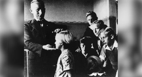 در این عکس، یک معلم آلمانی کودکی را که ویژگی‌های «آریایی» دارد، انتخاب، معرفی و از او ستایش می‌کند. هدف این بود که کودکان بیاموزند چه‌گونه نژاد‌ها را تشخیص دهند و درباره‌شان قضاوت کنند. آلمان، در دوران جنگ جهانی دوم
